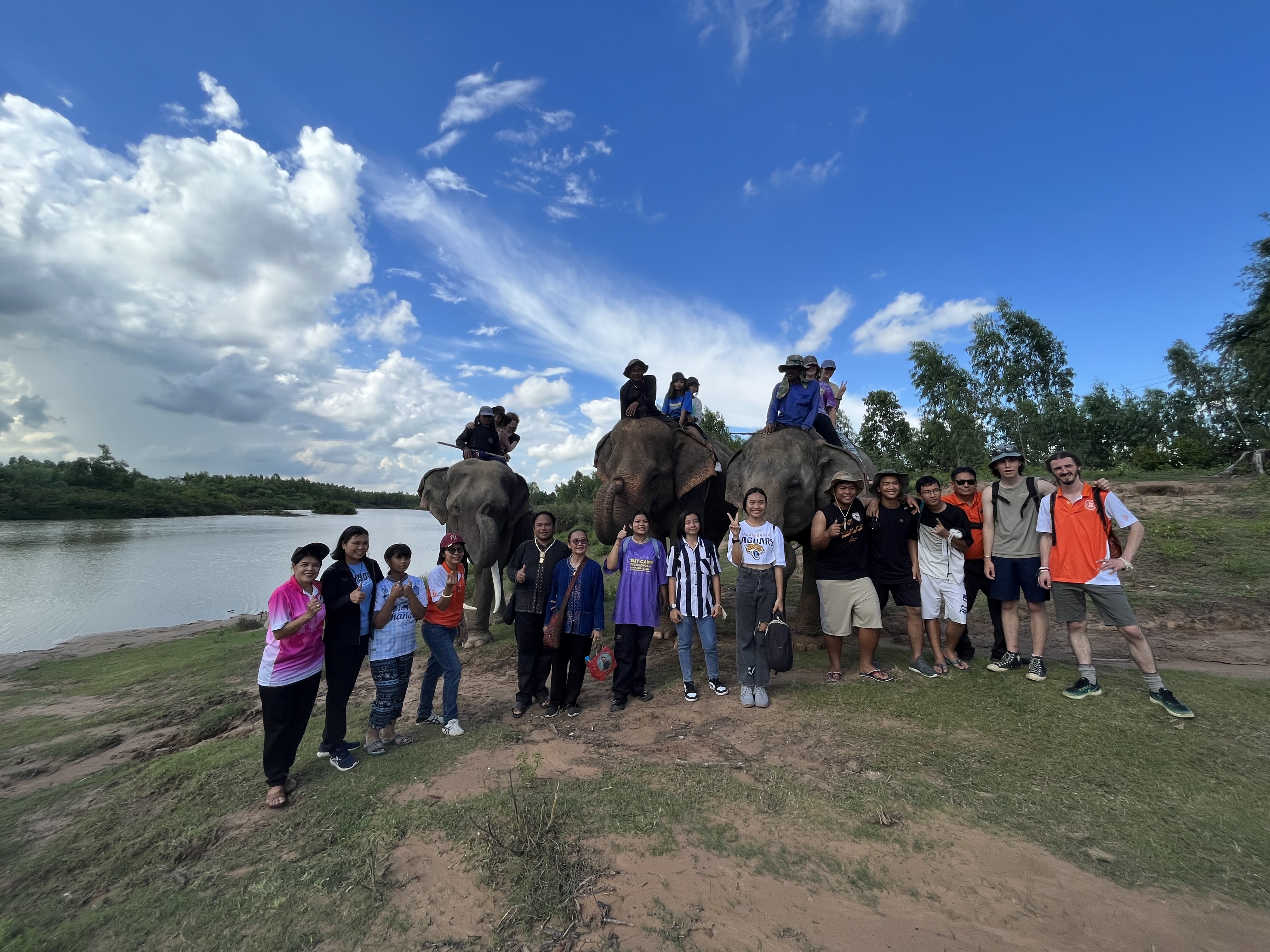 A group of Thai and American students smile together under a blue sky, some students are riding elephants.
