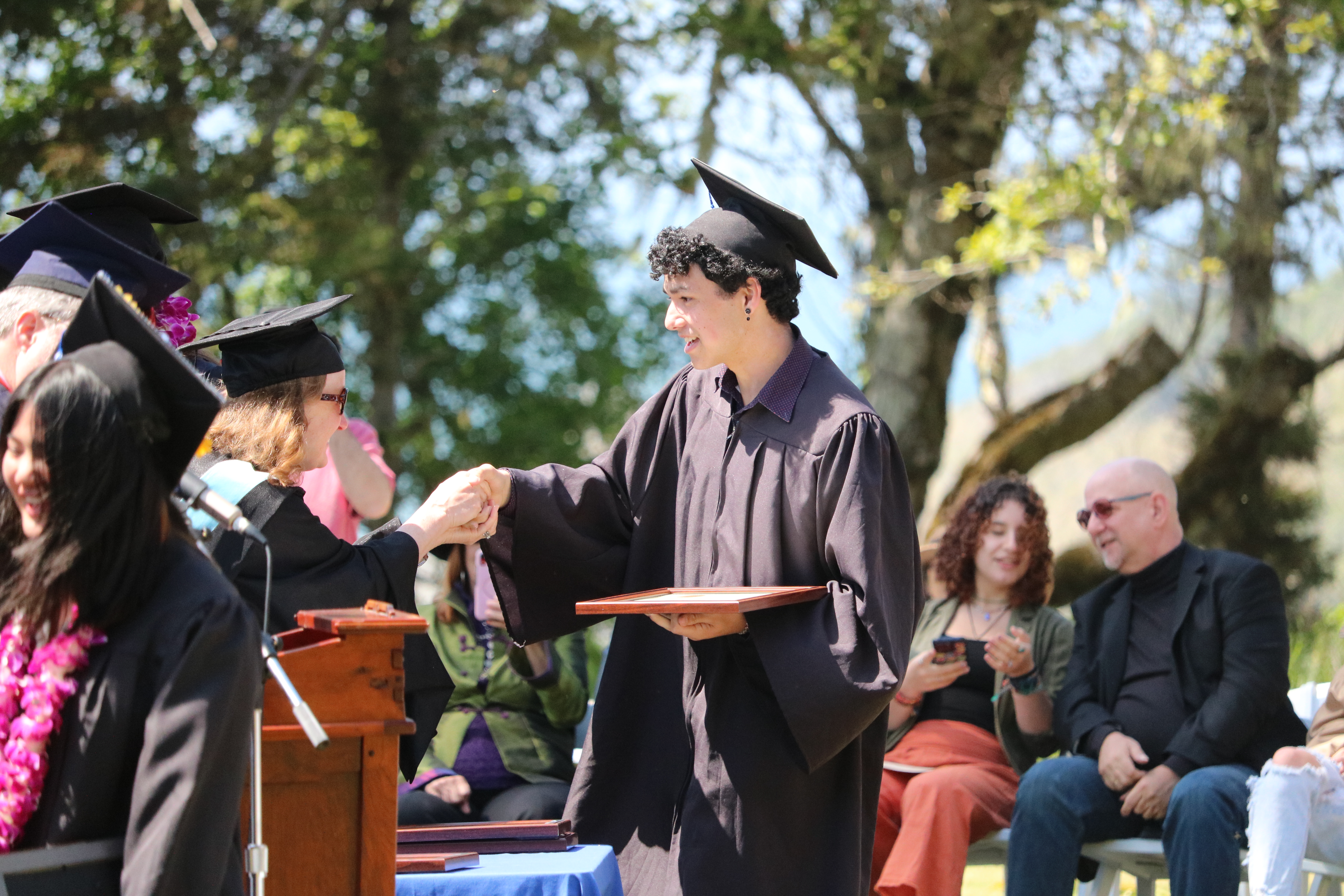 High school student in black graduation cap and gown shakes hands and receives his high school diploma at an outdoor graduation ceremony in Humboldt County, California..